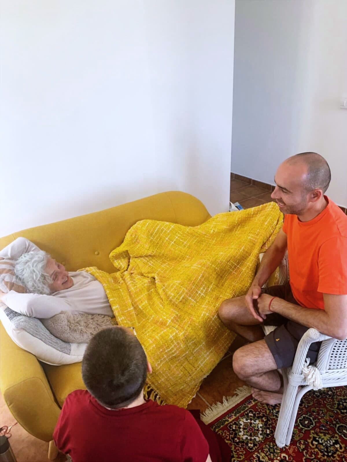 Una mujer mayor descansa en el sofá de su casa, tapada con una manta amarilla, junto a dos voluntarios